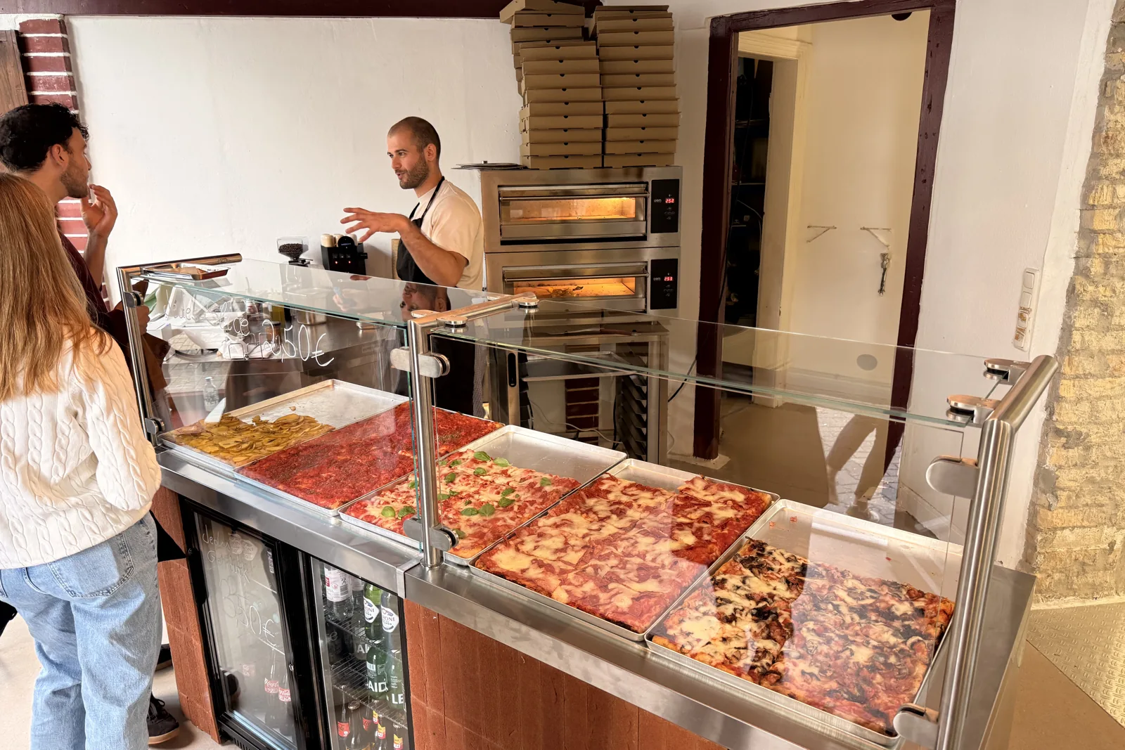 Inside SLICE CLUB: a guest at the counter, trays in the display, the pizzaiolo at the oven